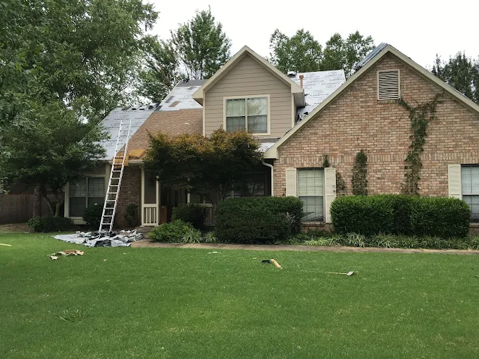 Two-story brick house with a ladder leaning against the roof and debris on the front lawn after storm damage
