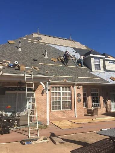 Roofing workers repairing a brick house roof under a clear blue sky, with ladders and materials on the patio.