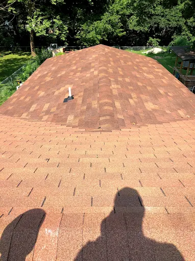 Brown shingle roof with a small vent, trees in the background, and a person’s shadow at the bottom edge
