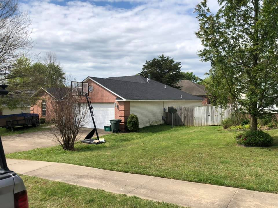 Suburban backyard with green lawn, trees, fence, driveway, and a brick house under a cloudy sky