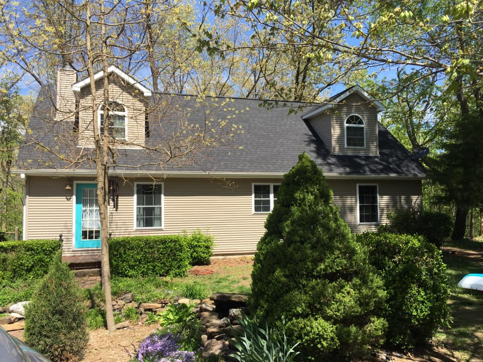 Suburban house with tan siding, dark roof, dormer windows, and a bright turquoise front door surrounded by shrubs