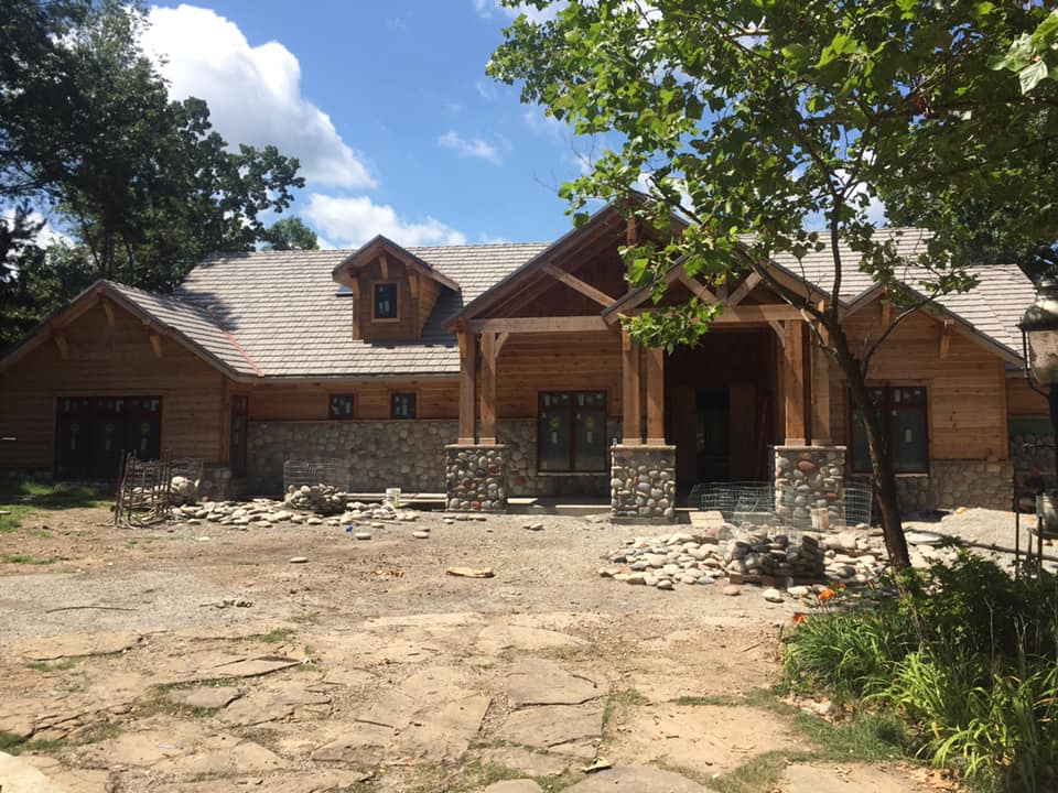 Log cabin-style house with stone pillars and a dirt yard under trees on a sunny day