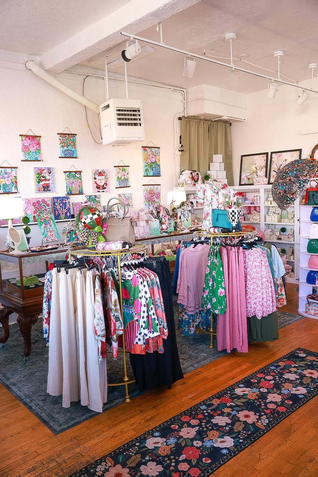 Colorful clothing racks in a boutique with floral artwork, white walls, and patterned rugs on wooden floors