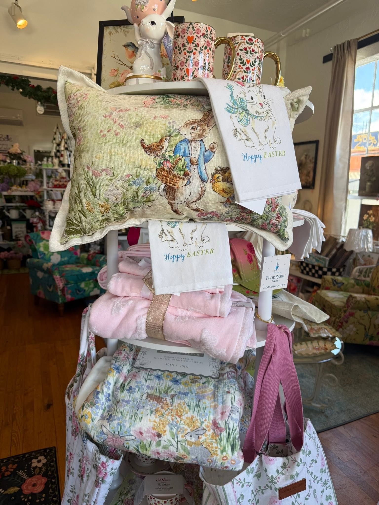 Display of decorative pillows and blankets in a shop, with floral and pastel patterns stacked on shelves.