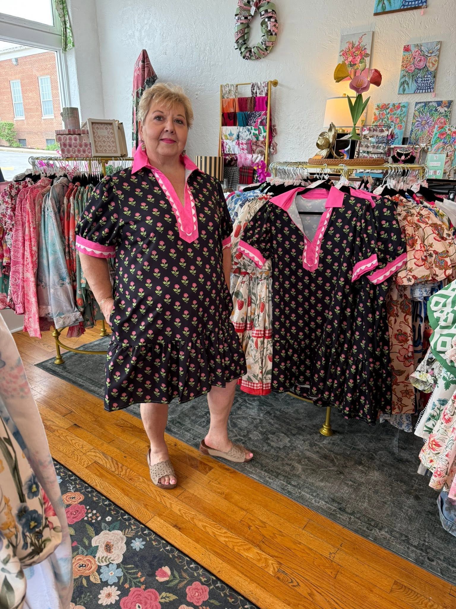 Woman in a boutique, standing beside racks of black dresses with bright pink trim.