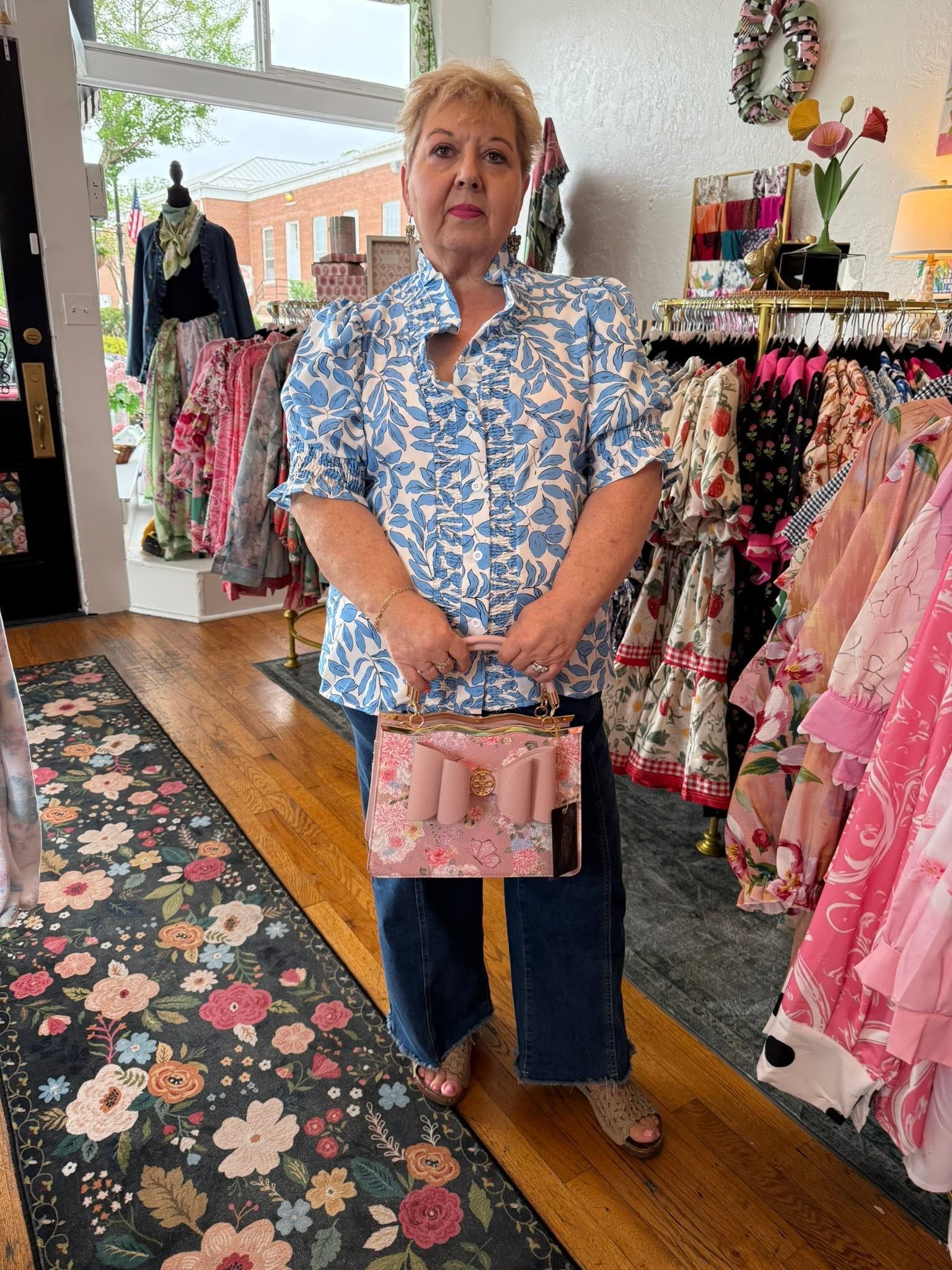 Woman standing in a boutique, holding a pink handbag, surrounded by colorful dresses and floral decor.