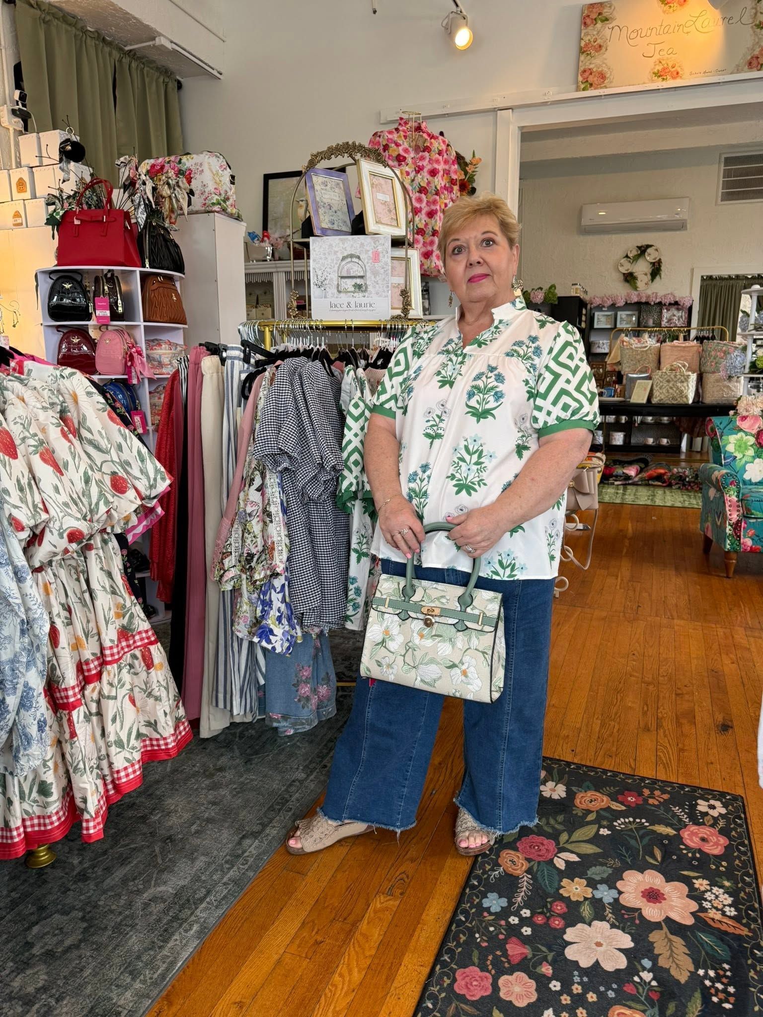 Woman standing in a floral clothing boutique, holding a handbag and facing the camera.