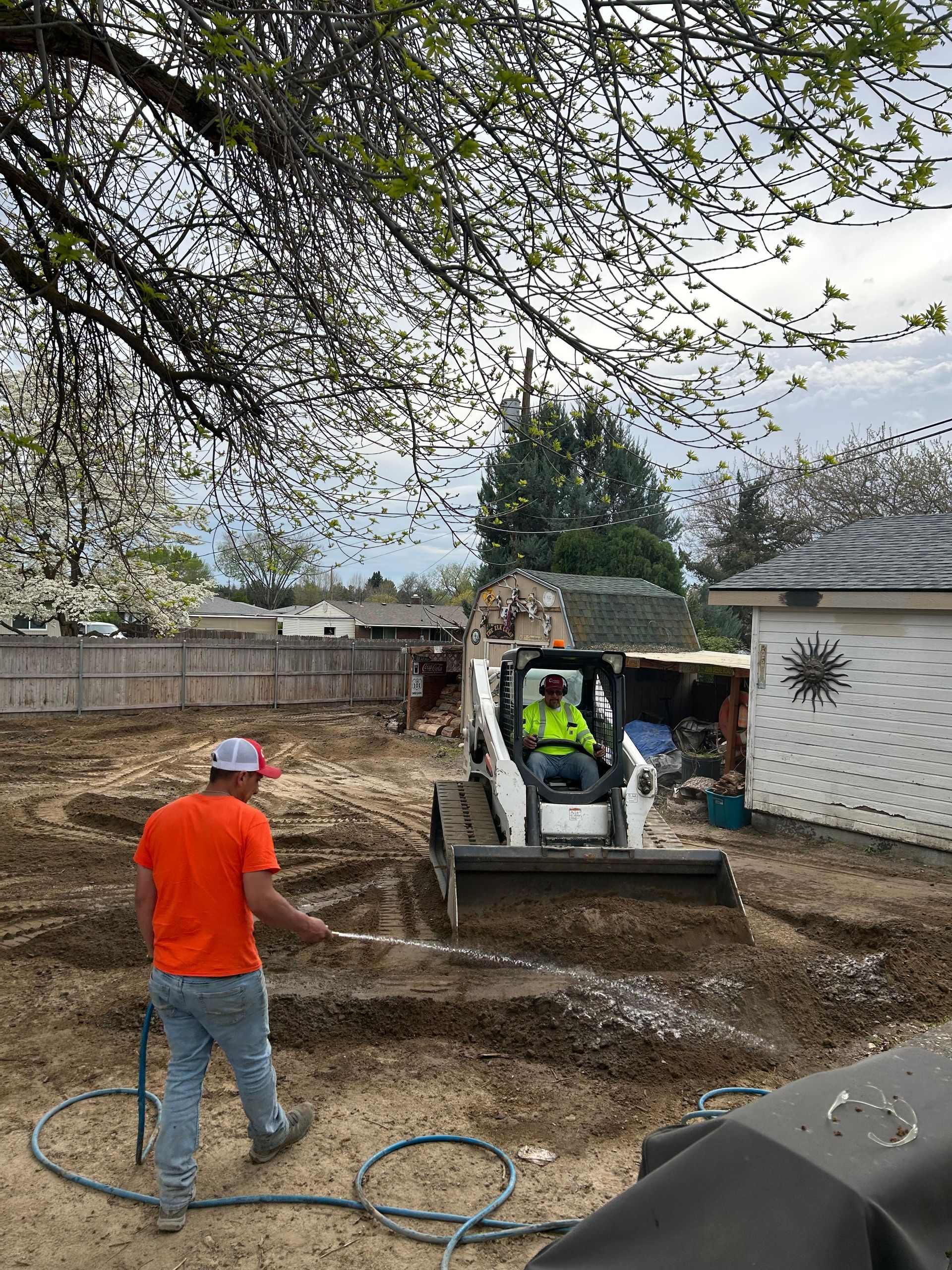 Worker in orange shirt guiding a skid steer in a dirt yard beside a house and wall.