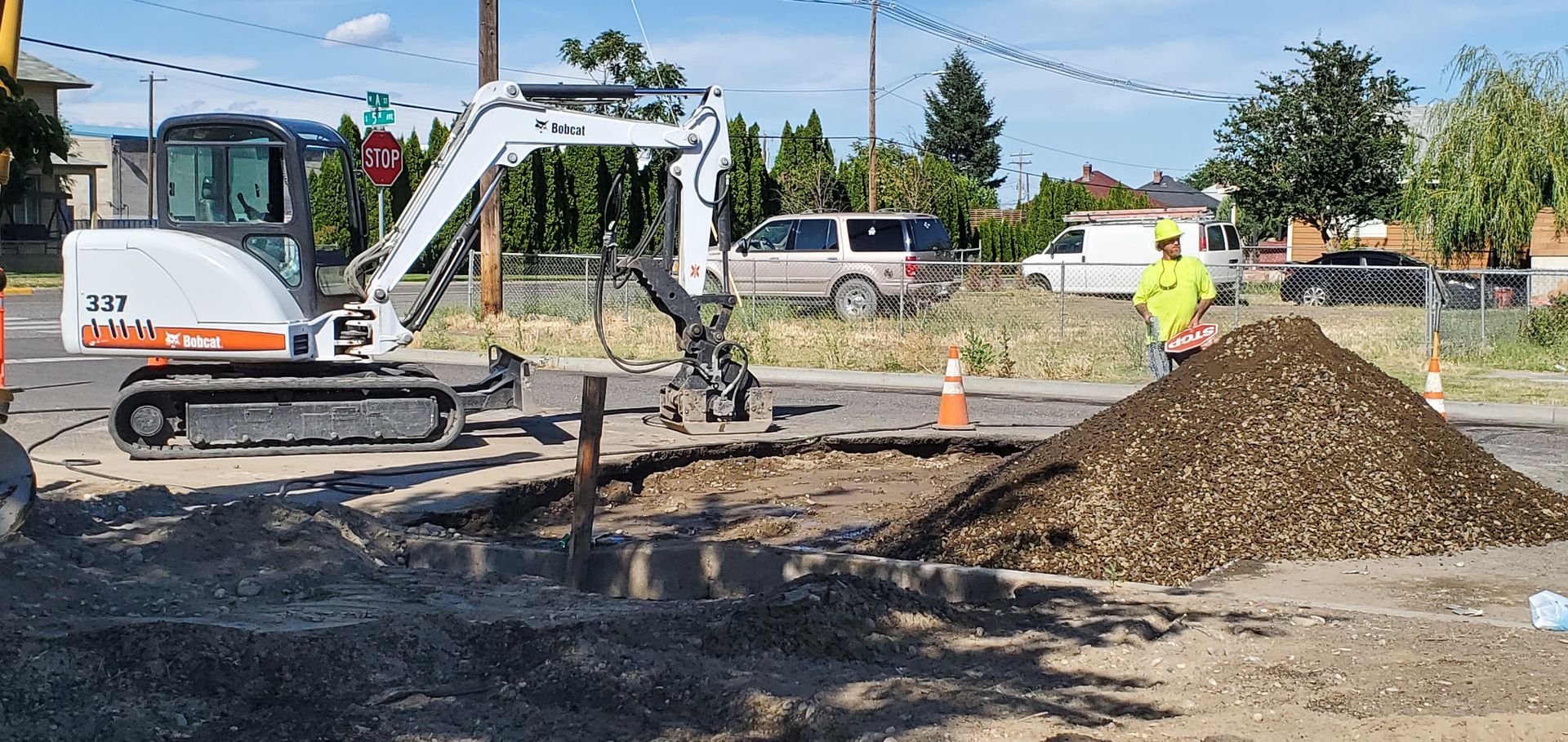 Excavator loading dirt at a roadwork site beside a large pile of gravel and orange cones.