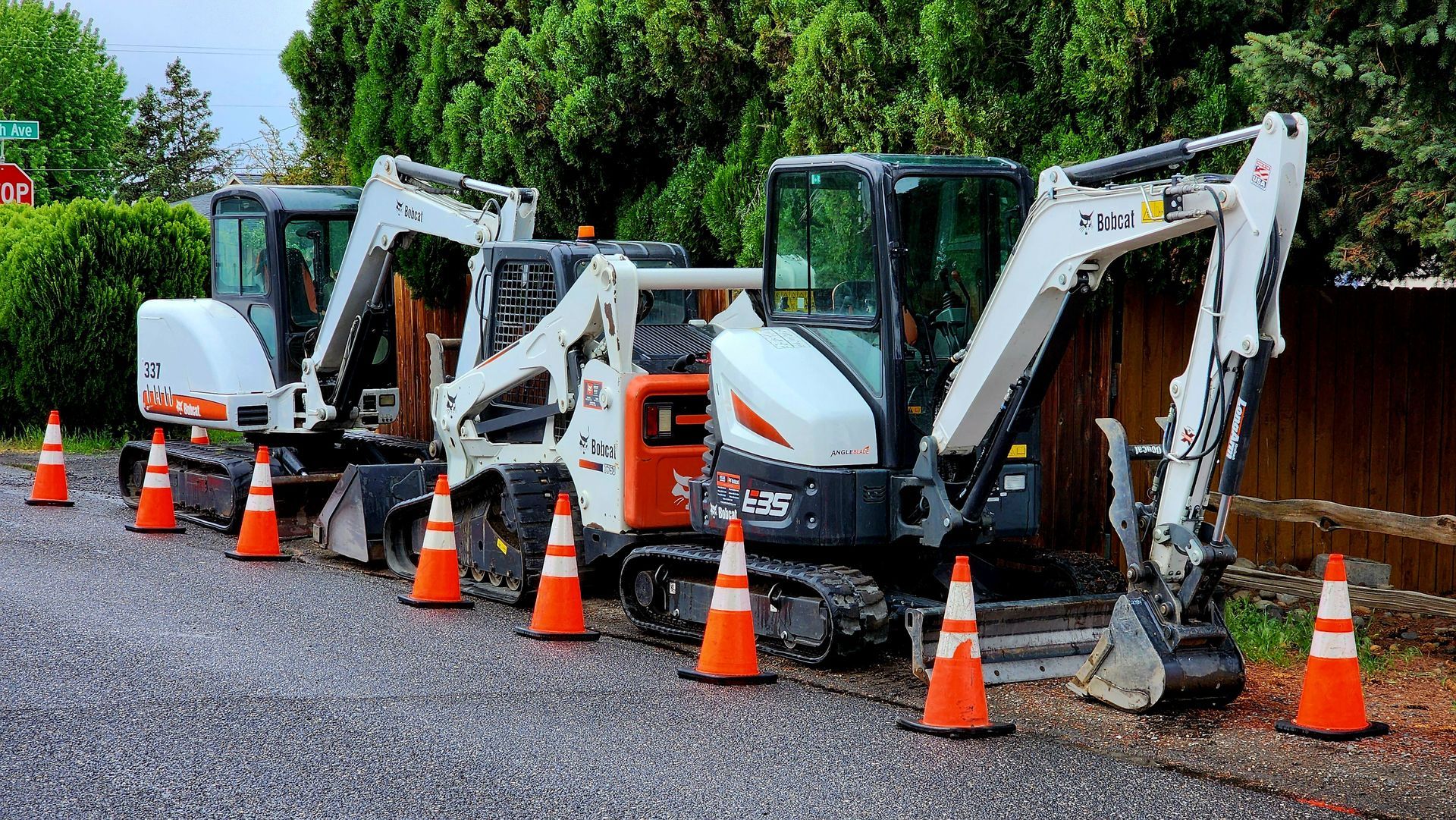 Three mini excavators lined up behind orange cones on a gravel work site