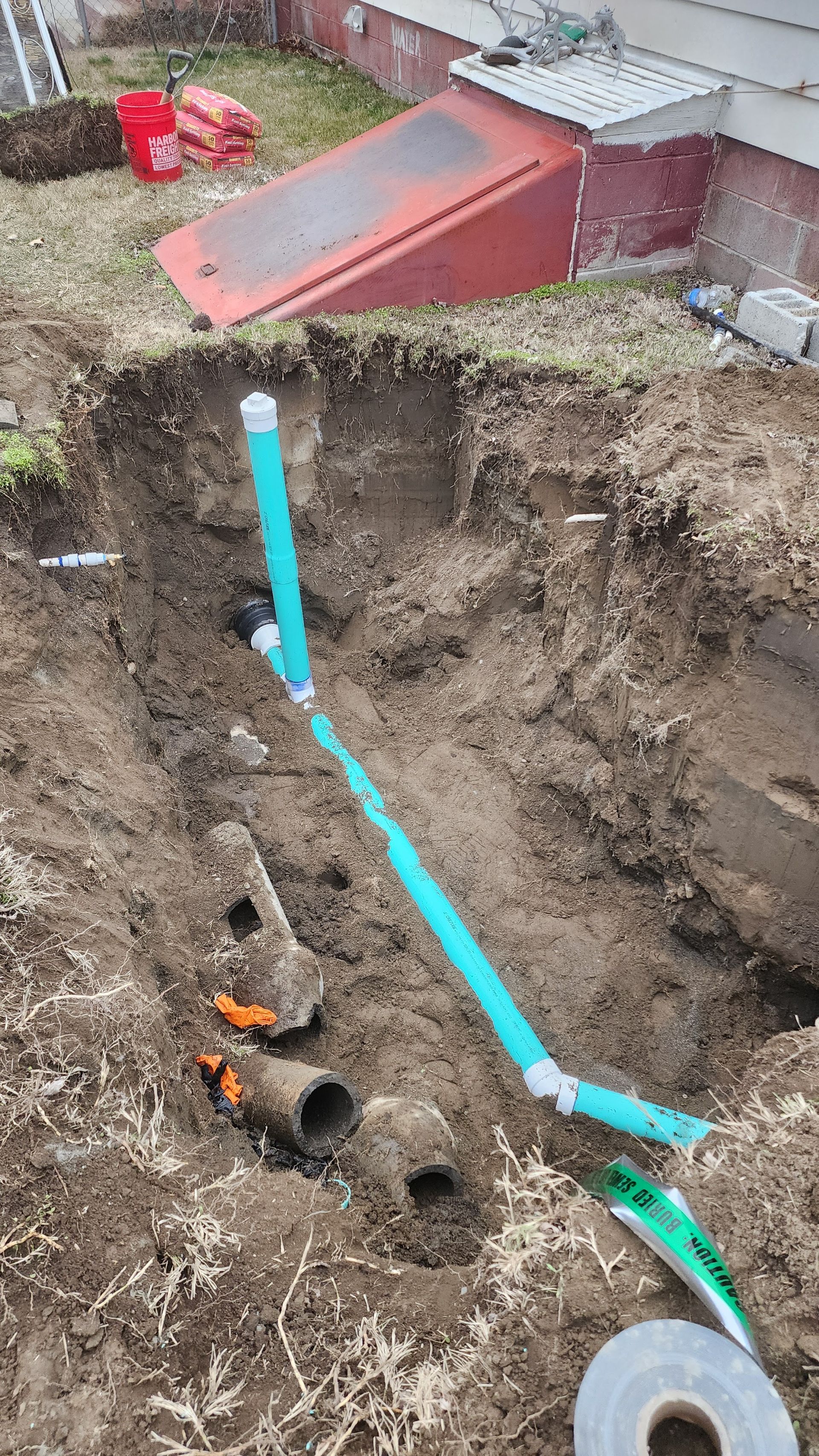 Backyard trench excavation with a blue pipe exposed and soil piled beside the house