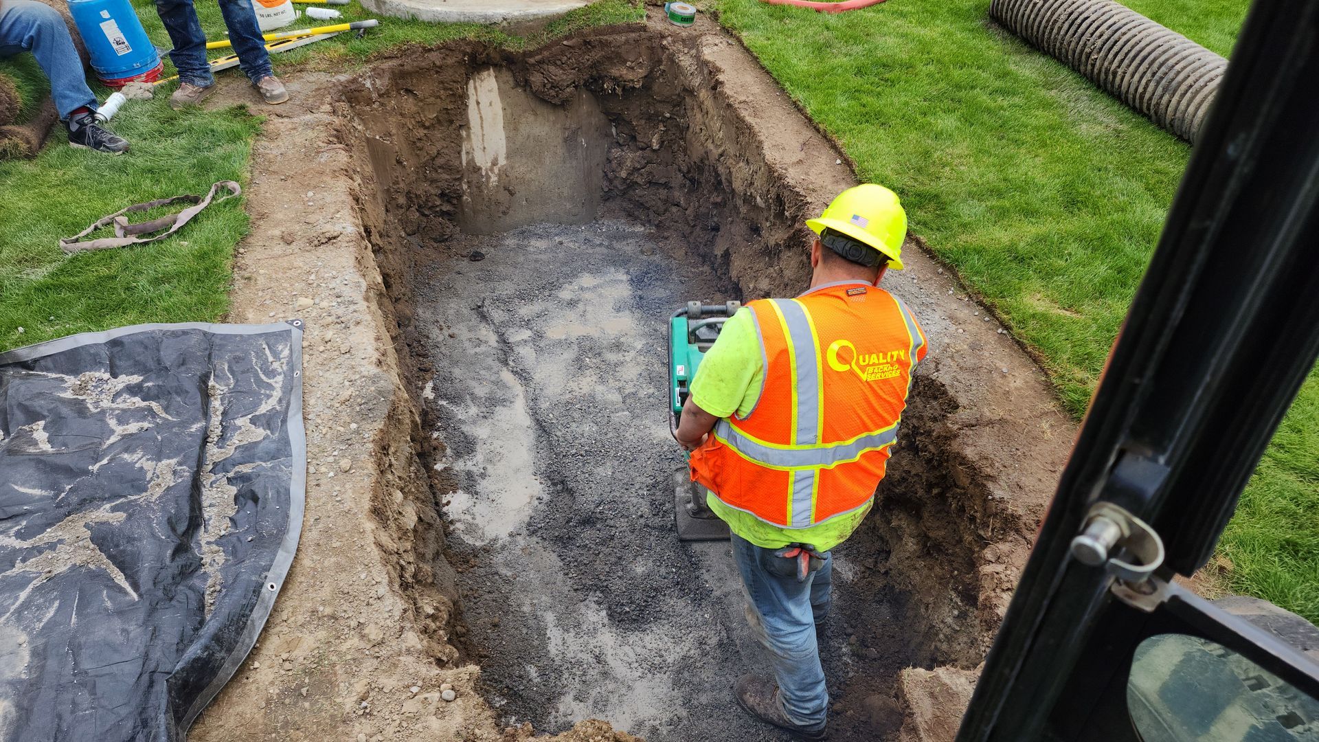 Worker in orange safety vest and hard hat inspecting a deep excavation trench on a grassy site