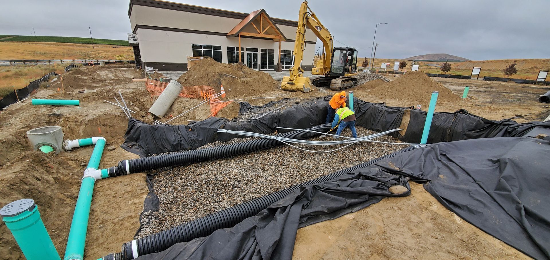 Construction site with drainage pipes, black geotextile, and a worker near a house foundation.