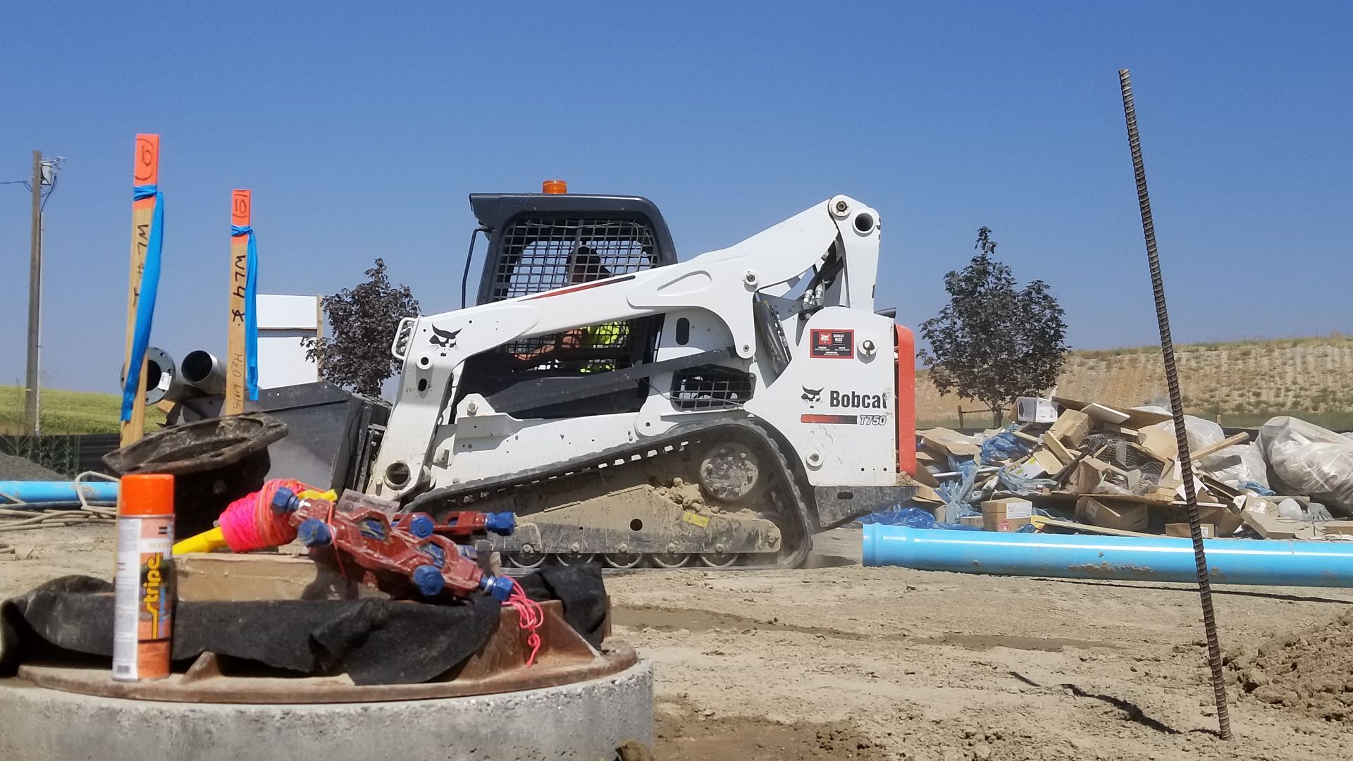 White skid-steer loader dumping dirt at a construction site beside a blue pipe and orange safety cones