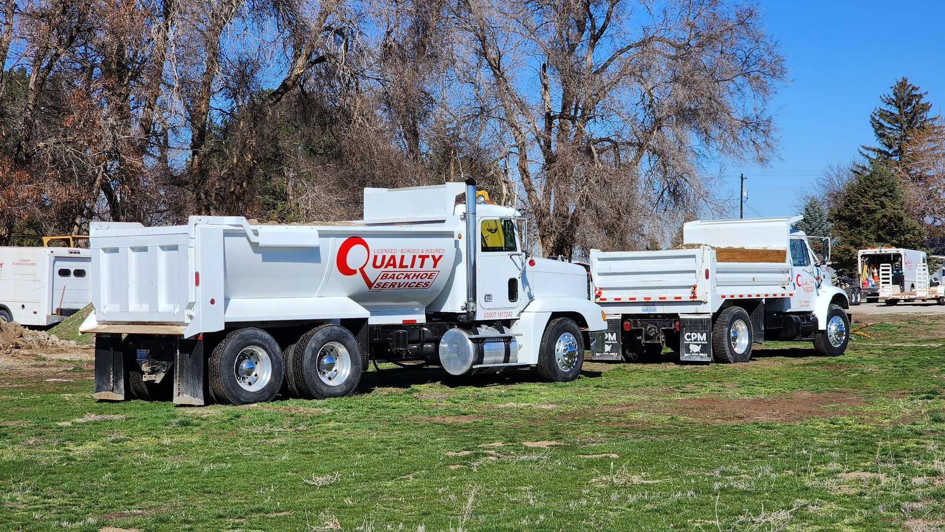 Two white dump trucks parked on a grassy lot under clear blue sky