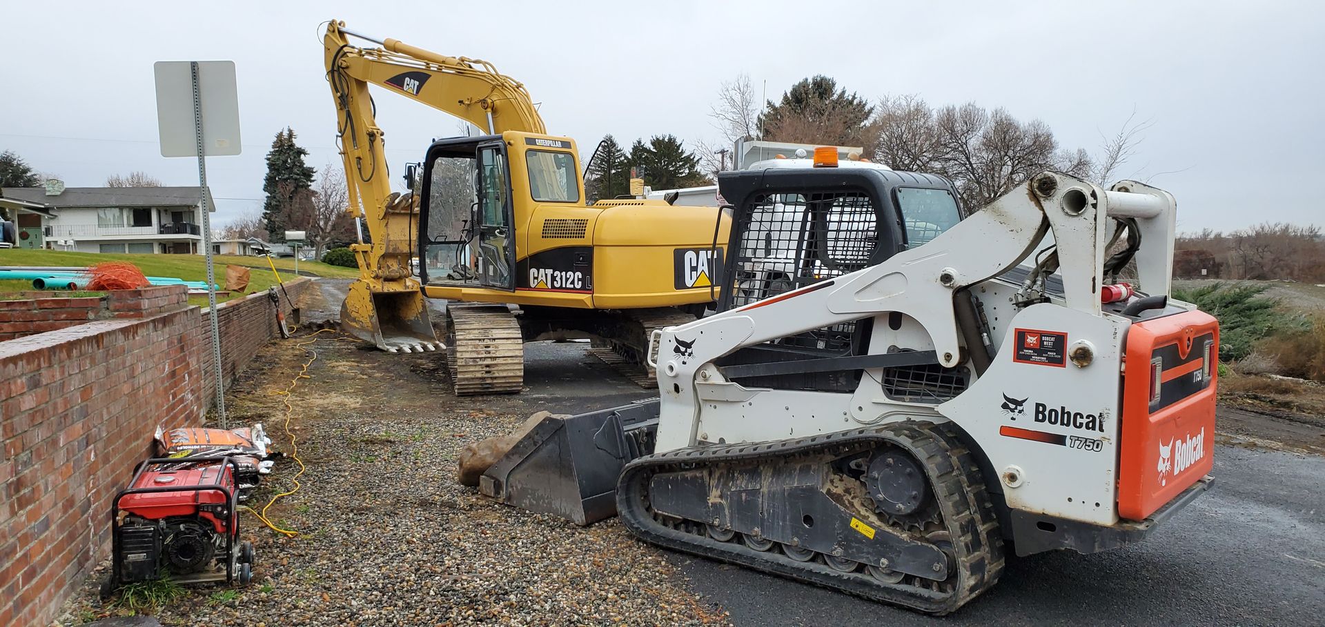 Yellow excavator and white track loader on a gravel lot near a brick wall and house.
