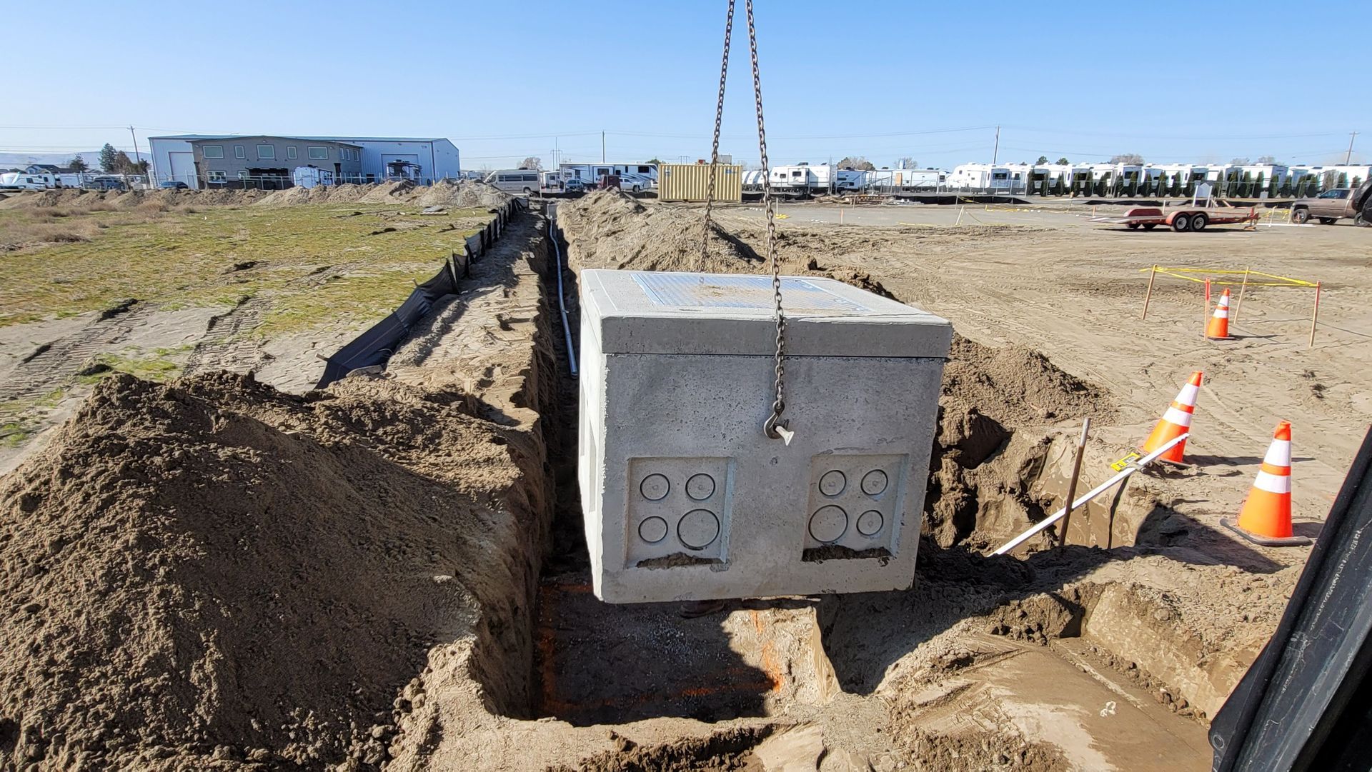 Concrete utility box being lifted by a crane at a construction site, with dirt trenches and orange cones nearby
