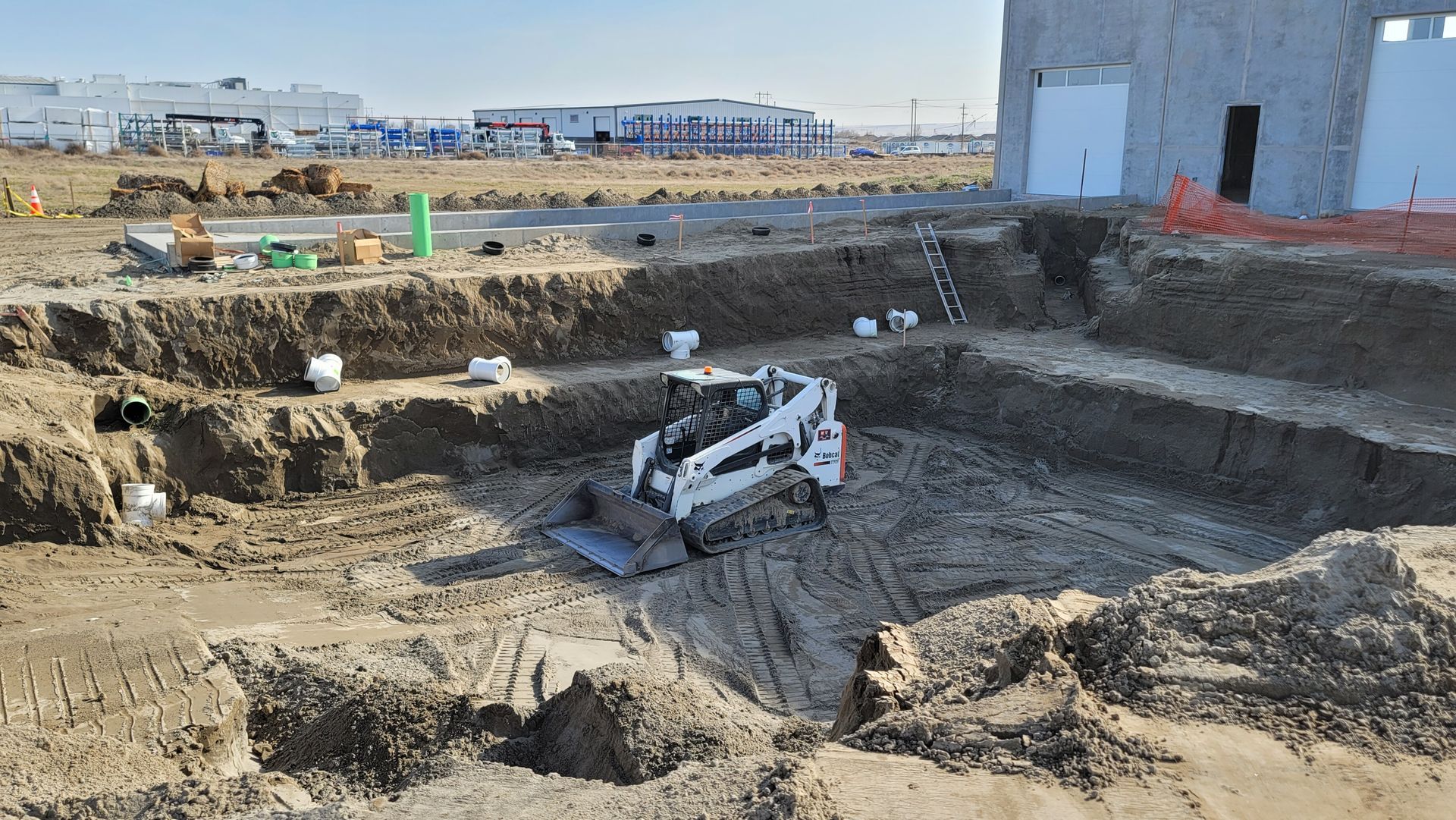 Excavator digging a large construction pit beside a building on a dirt site.