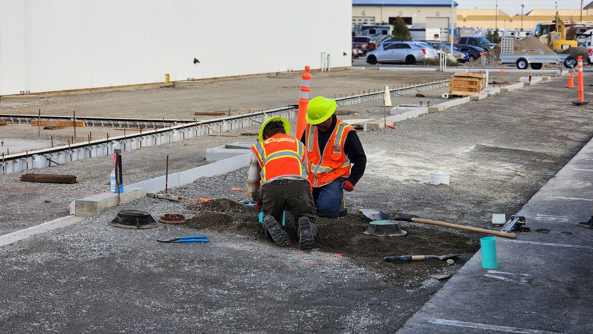 Two construction workers in orange safety vests kneel at a roadwork site near rebar and gravel.