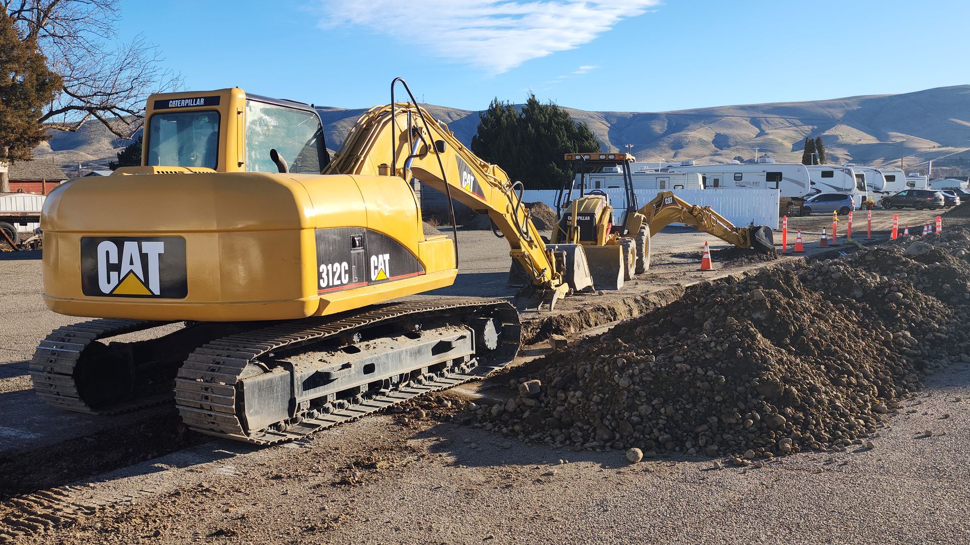 Yellow CAT excavators working on a roadside dirt pile under a clear blue sky
