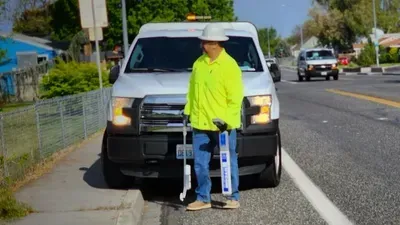 Person in neon yellow jacket standing on a sidewalk beside a white truck on a suburban street.