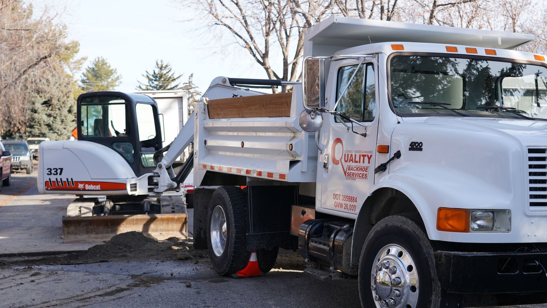 White dump truck beside a small excavator on a muddy roadside with trees in the background