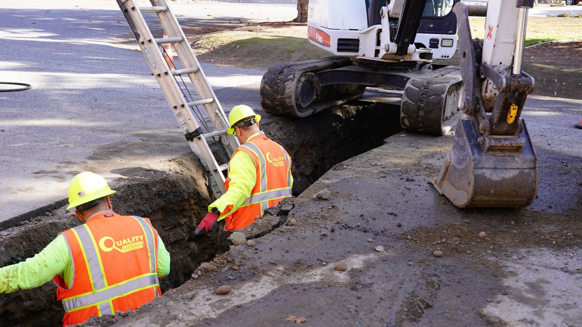 Construction workers in orange vests repair a road trench beside a backhoe and truck.