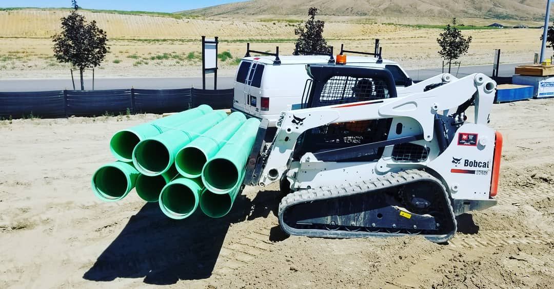 White skid-steer loader carrying bundled green pipes on a dirt lot under a bright sky