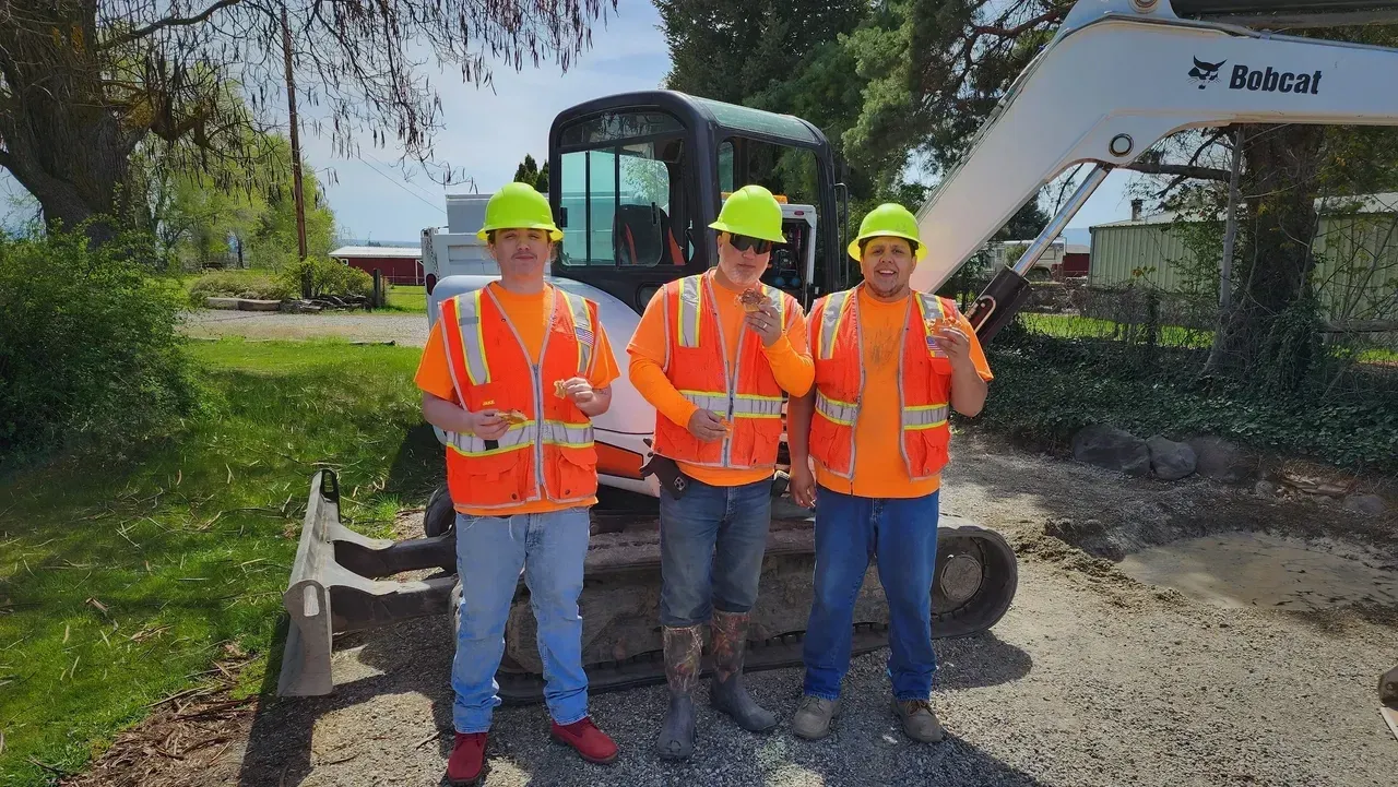 Three construction workers in orange vests and hard hats stand beside a concrete pump truck at a job site.