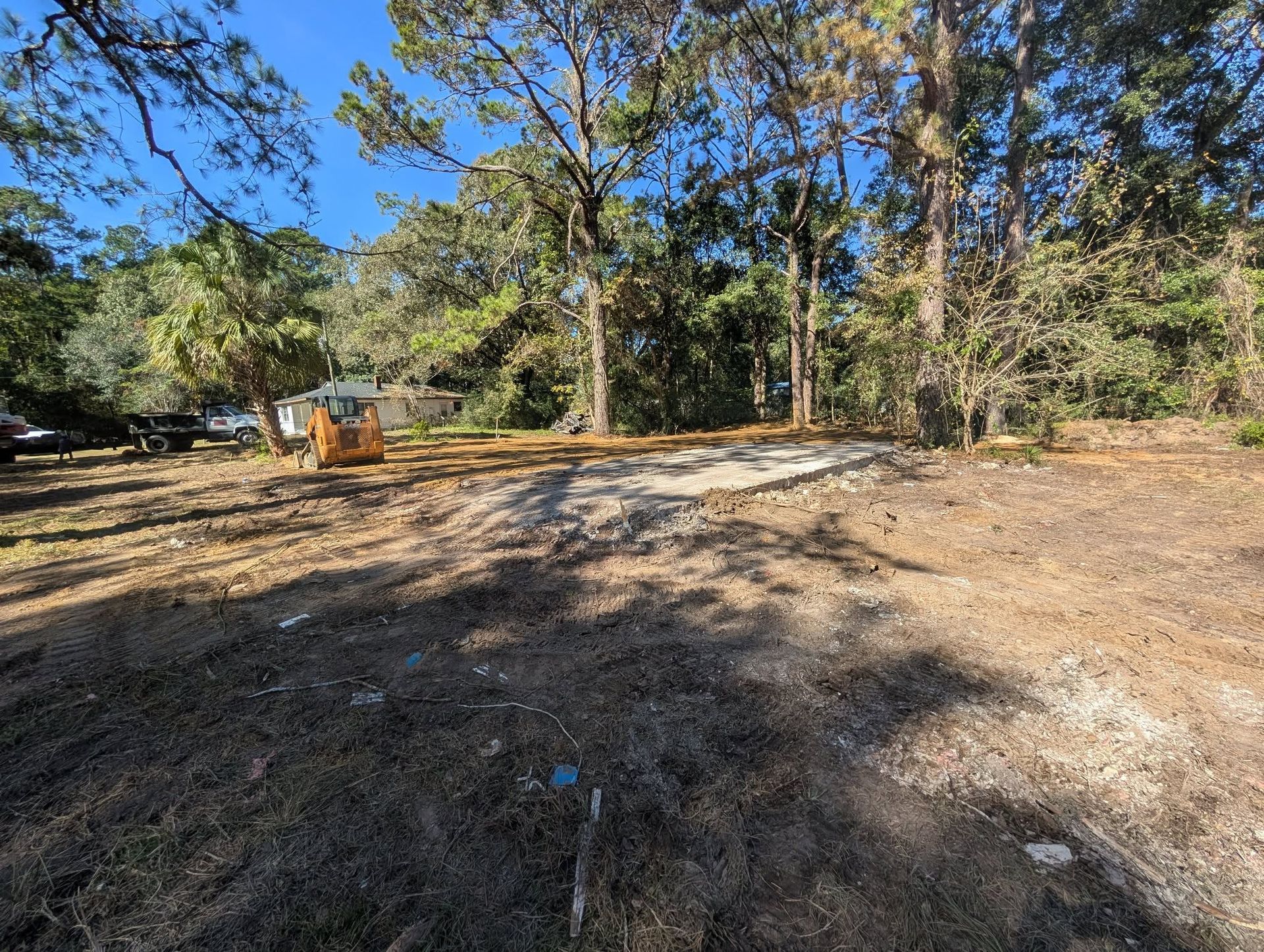 Wide dirt clearing with a small orange excavator, trees, and a blue sky on a sunny day