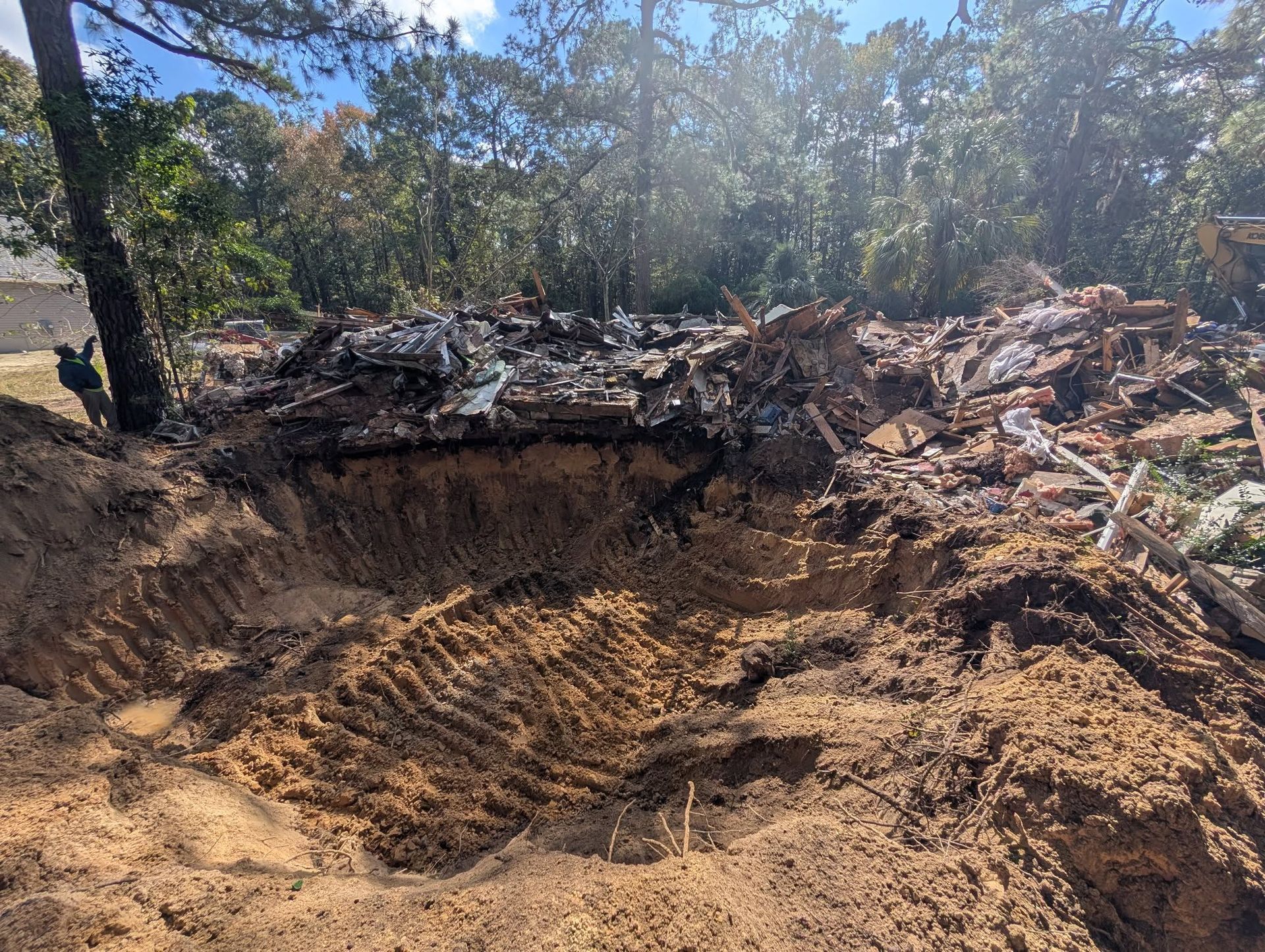 Large dirt excavation pit with a steep edge, debris piles, and trees under a sunny sky