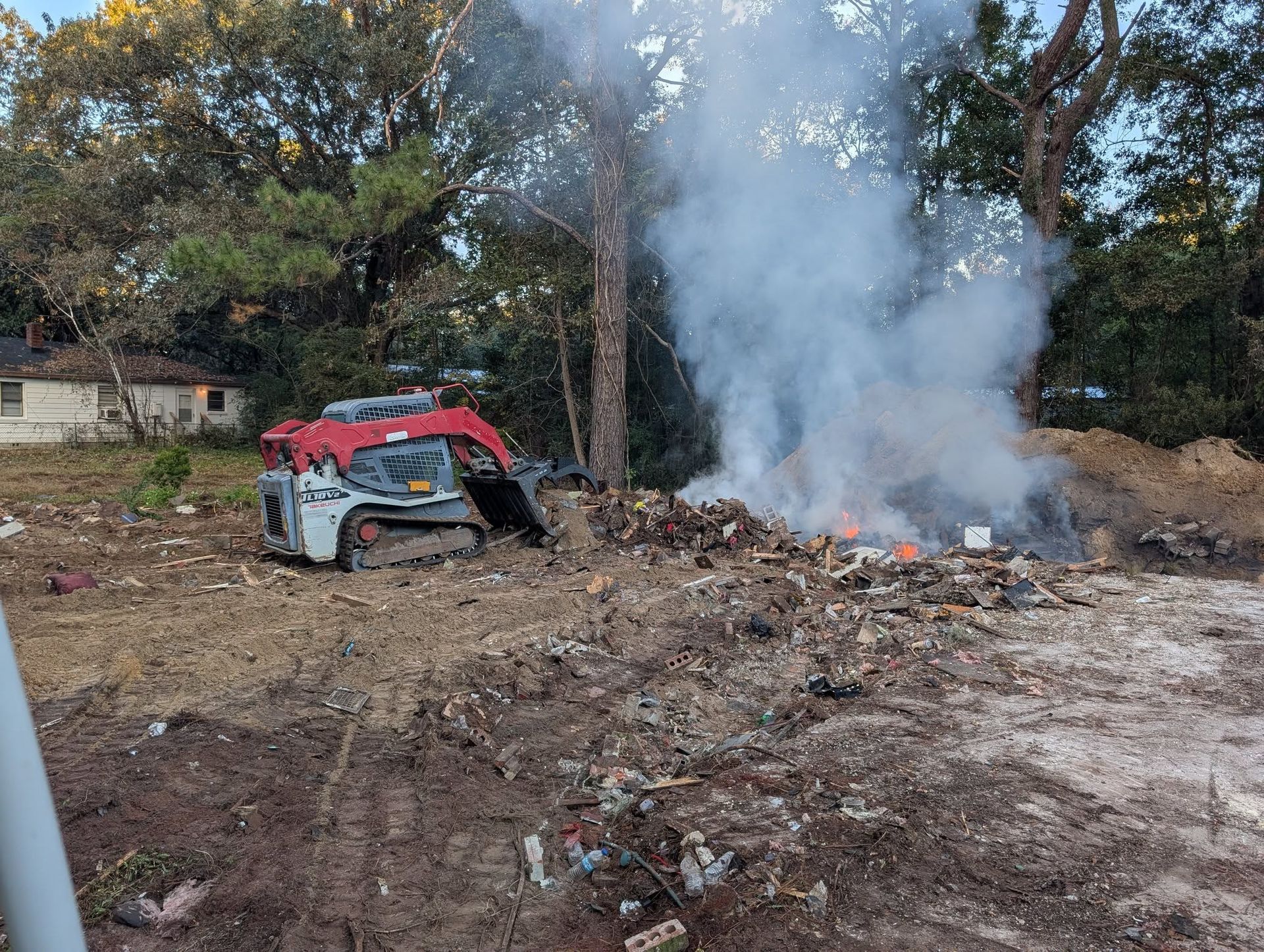 Burning debris in a dirt lot with a red vehicle nearby and smoke rising among trees