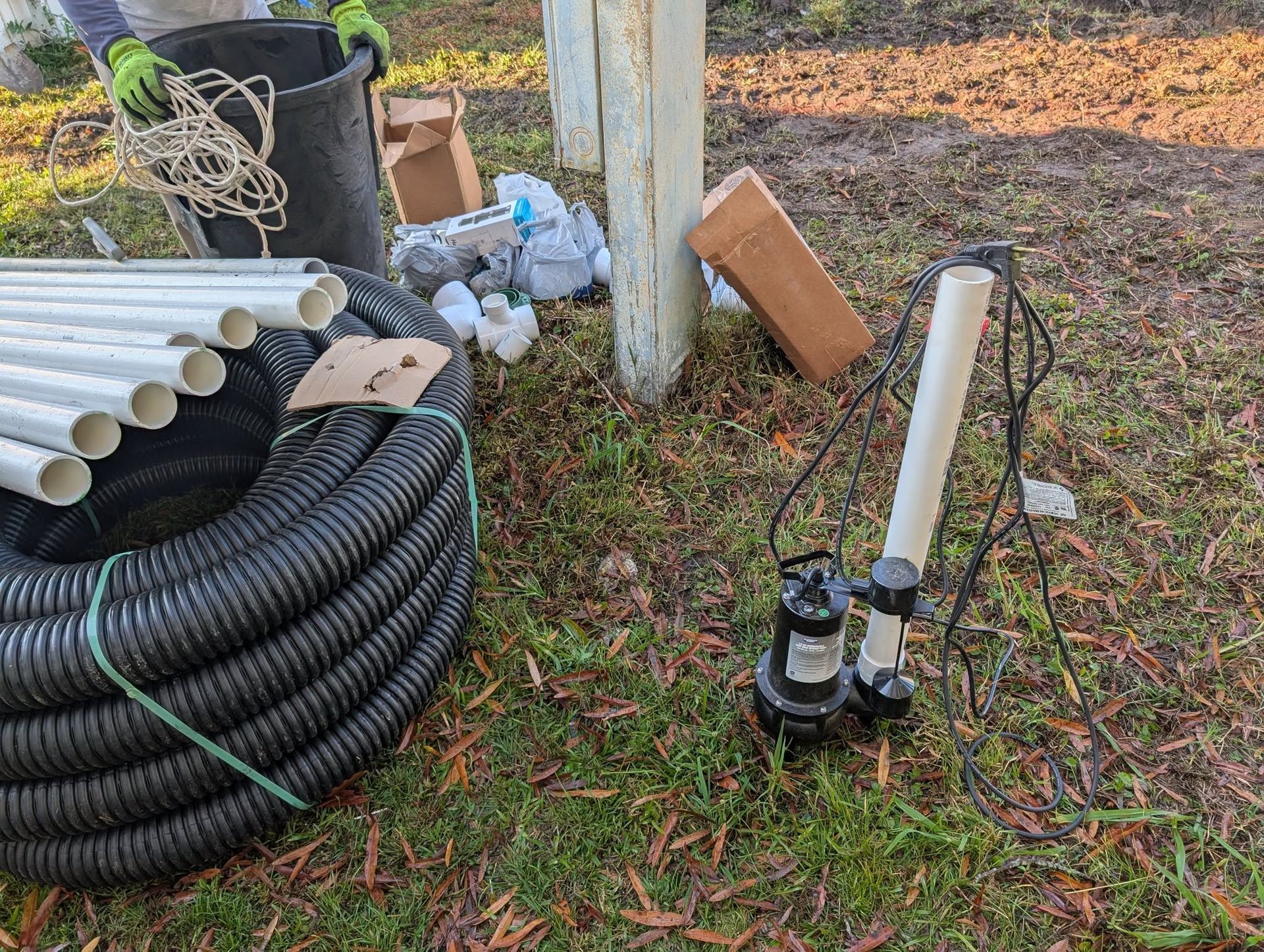 Outdoor utility setup with coiled black pipe, white PVC pipe, pump, and scattered tools on grass