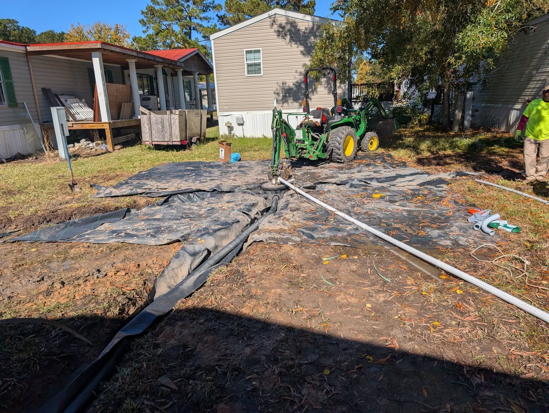 Yard with black tarp on ground, white pipes, and a small house with green furniture in the background