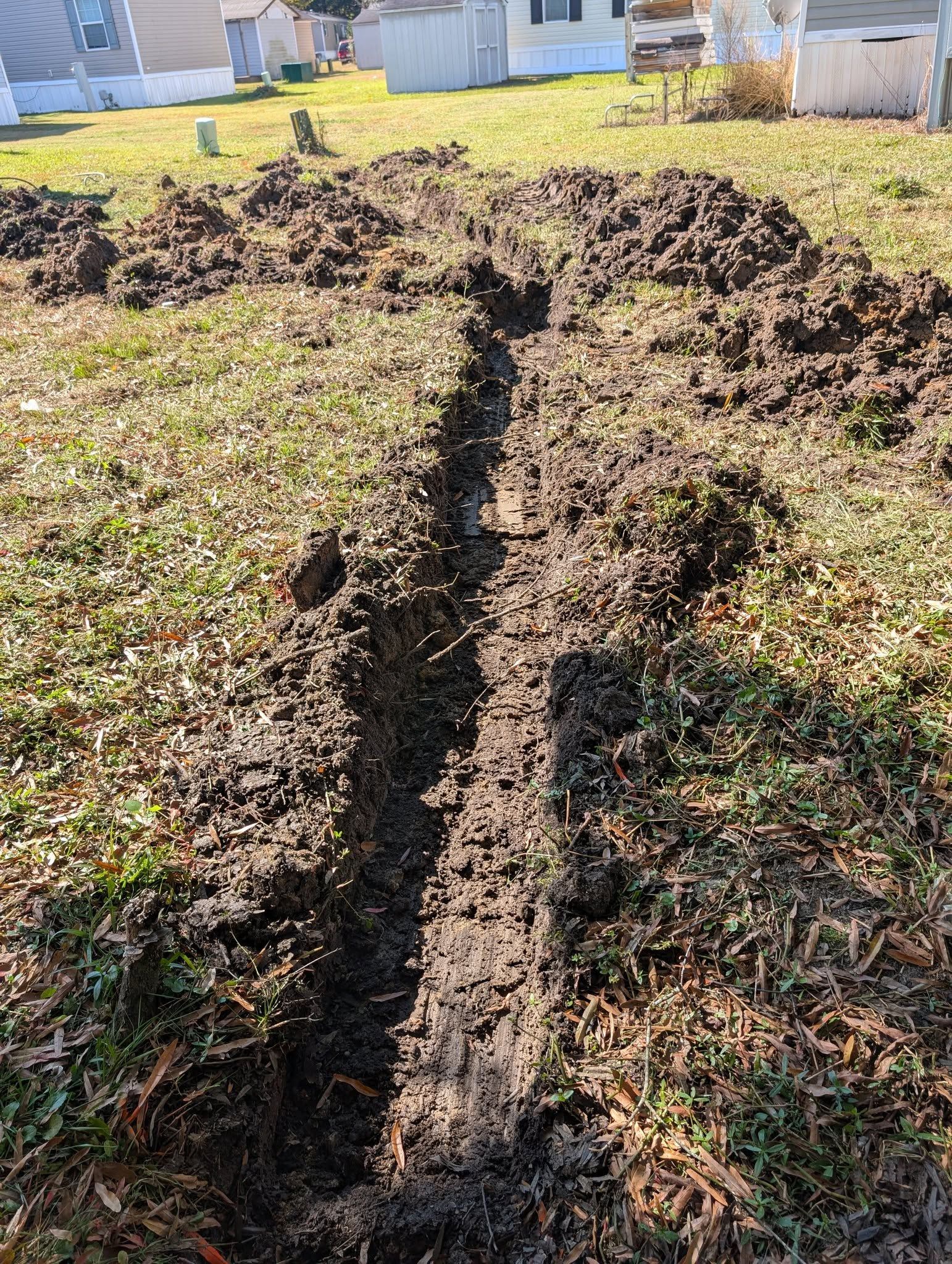 A muddy trench cutting through a grassy yard beside a house in bright daylight