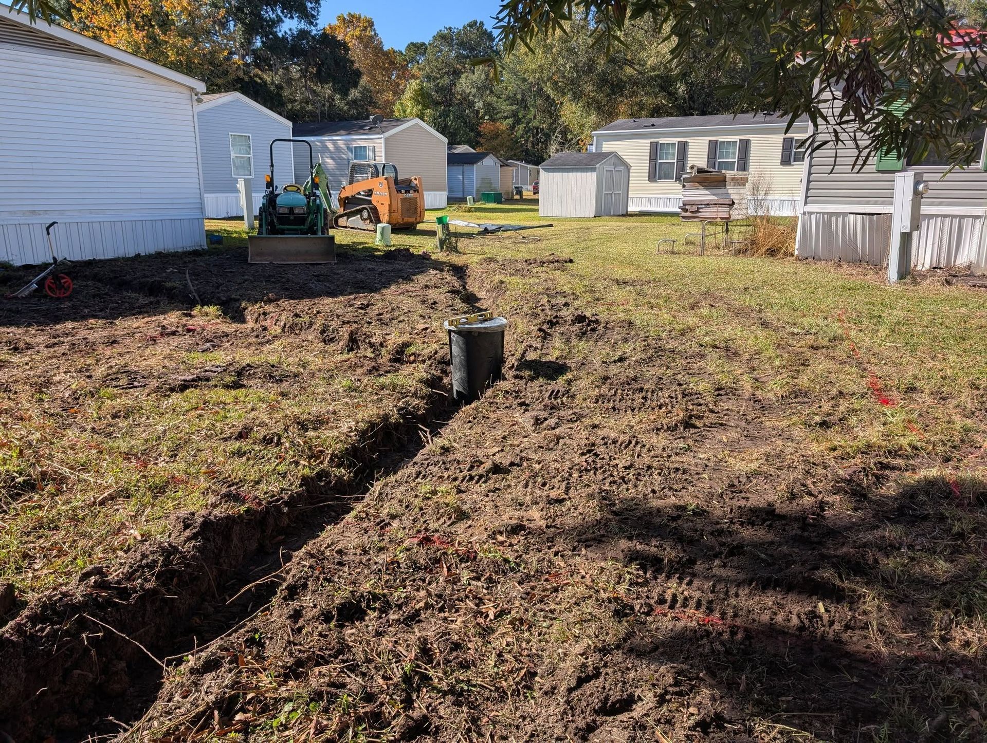 Backyard with a narrow trench, tree stump, white buildings, and an excavator in the distance.