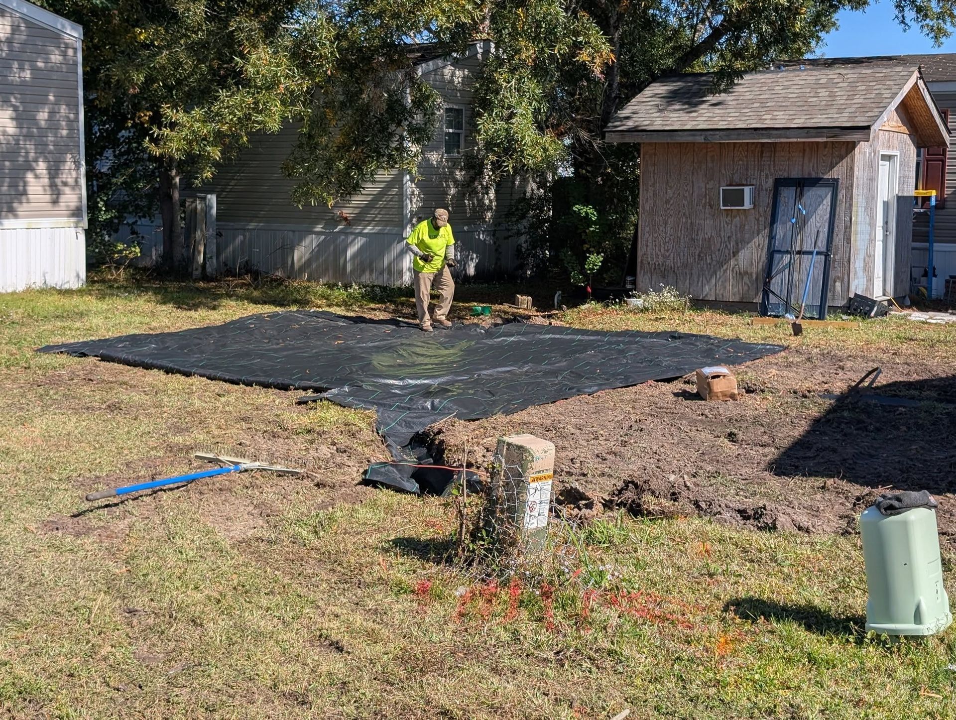 Worker laying black tarp in a yard beside a small shed and trees