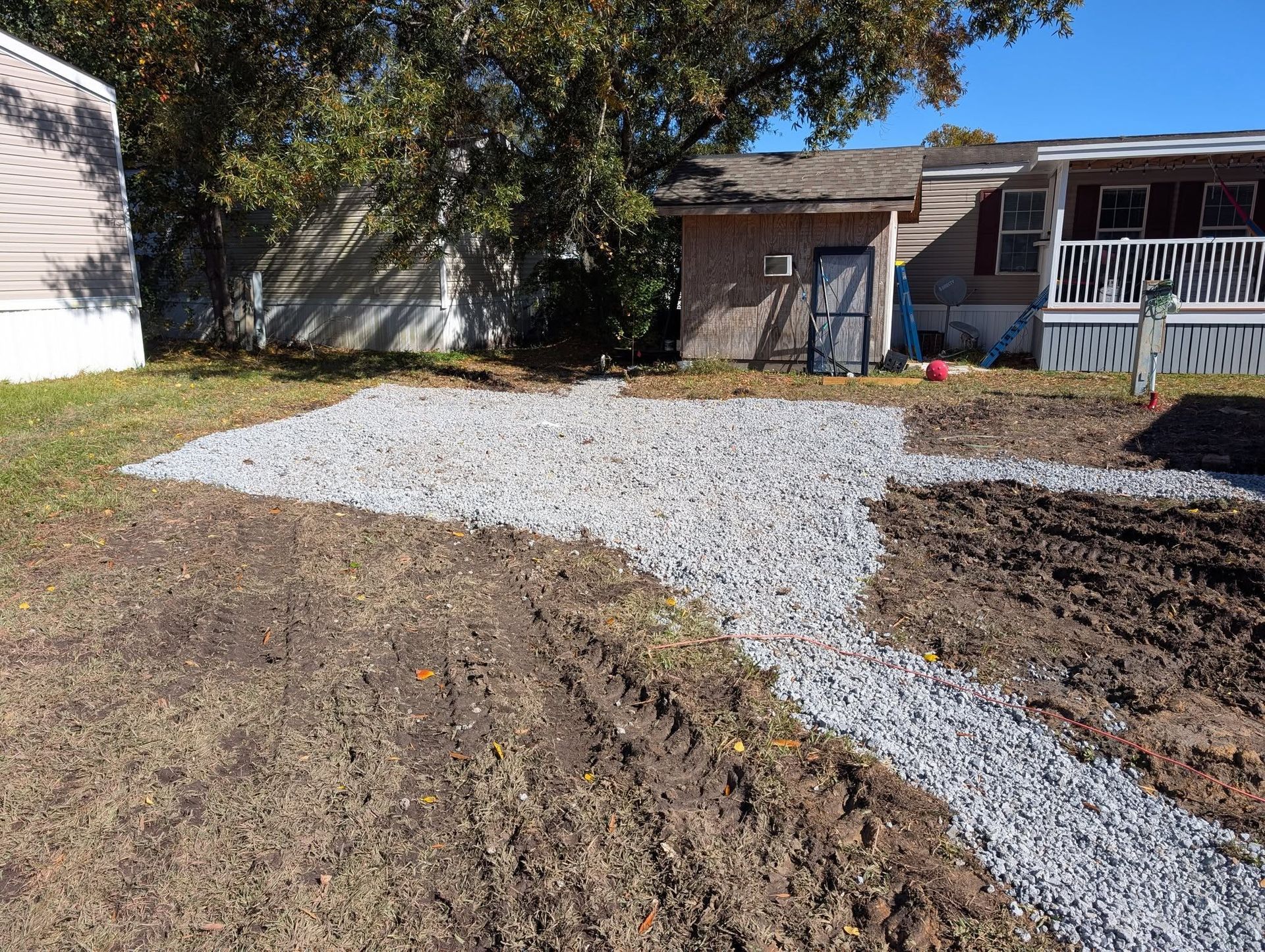 Gravel driveway under construction beside a house, with bare soil and a small shed in the background