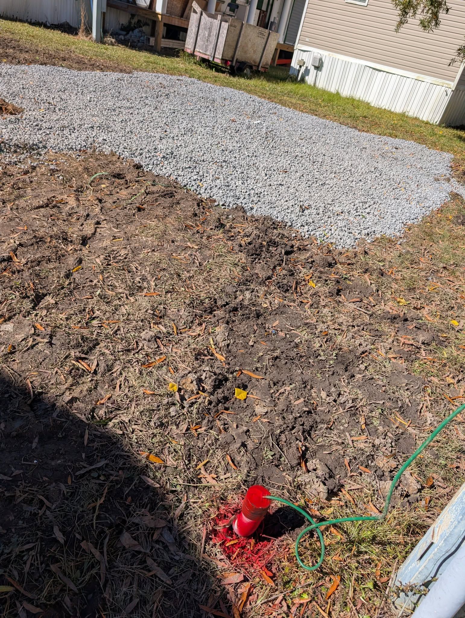 Rocky yard with a white gravel path, green hose, and red sprinkler near a house fence.