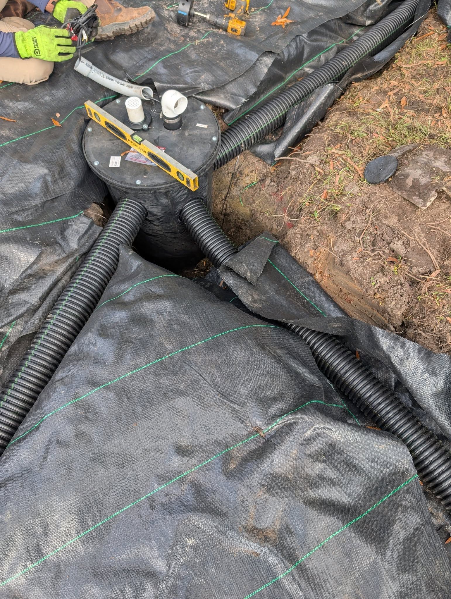 Black drainage pipe crossing a trench in a construction site, with dirt, plastic sheeting, and tools nearby