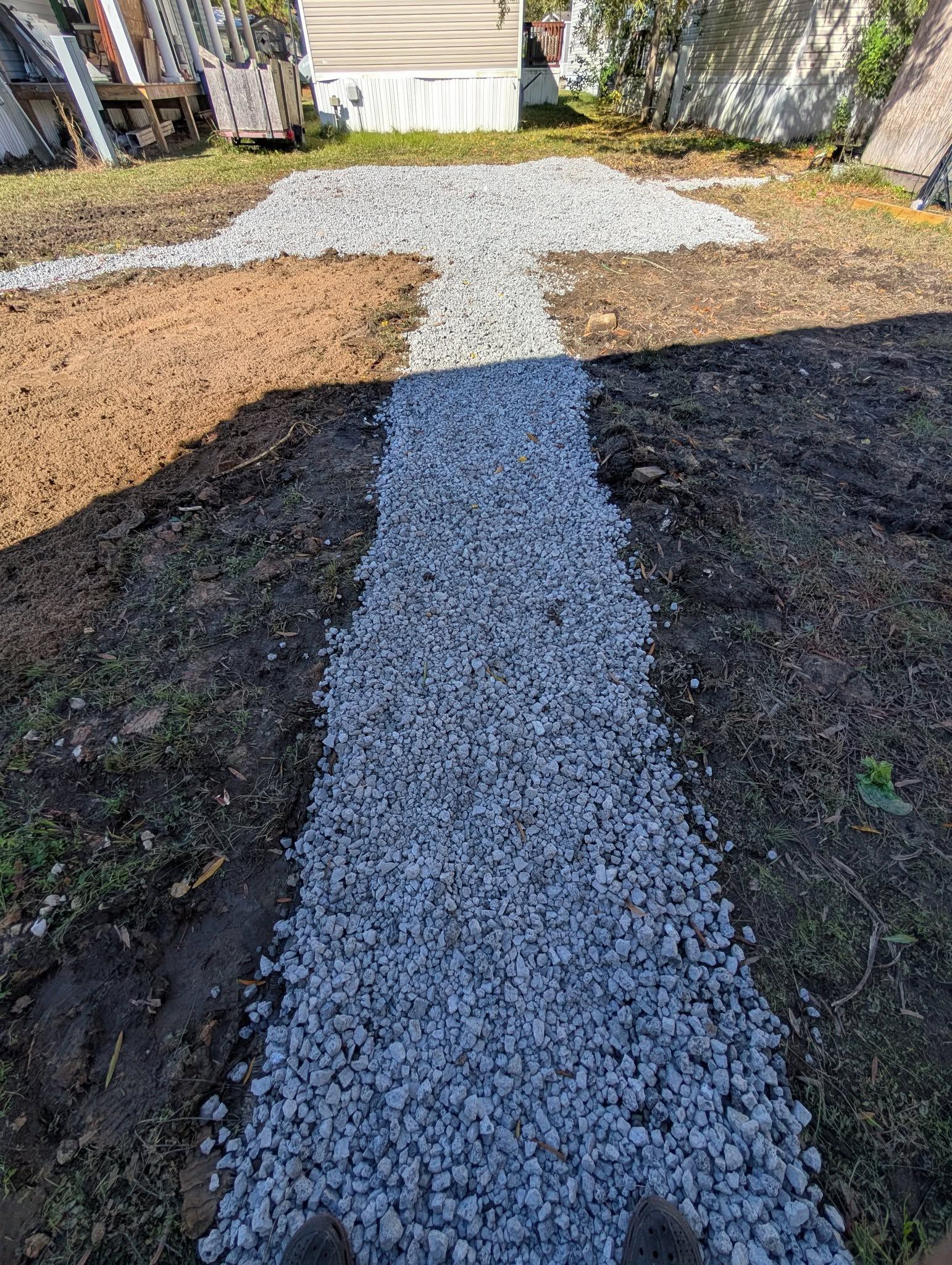 Gravel path through a yard, with trees and fencing on both sides, lit by sunlight and shadow.