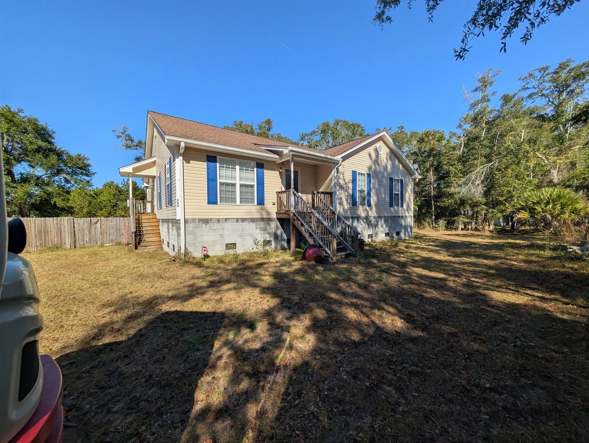 Small beige house with blue shutters on a sunny, wooded lot with dry grass and a driveway edge.