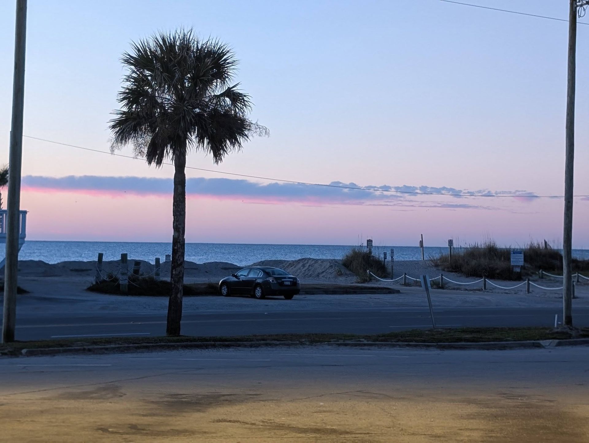 Palm trees and parked cars along a beach road at dusk, with pastel sky and calm ocean beyond.