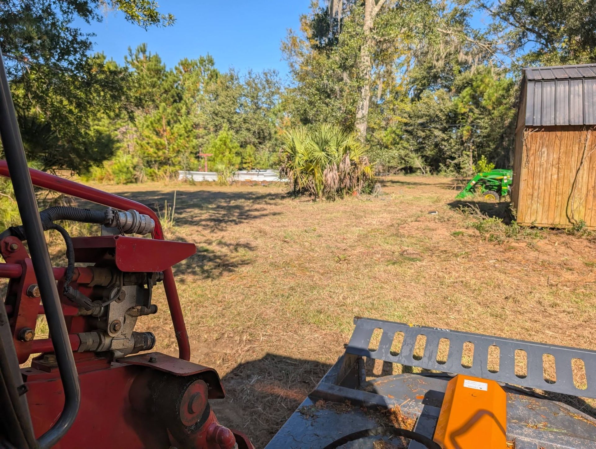 Sunlit yard with trees, a woodpile, red tractor parts, and a wooden shed on the right.