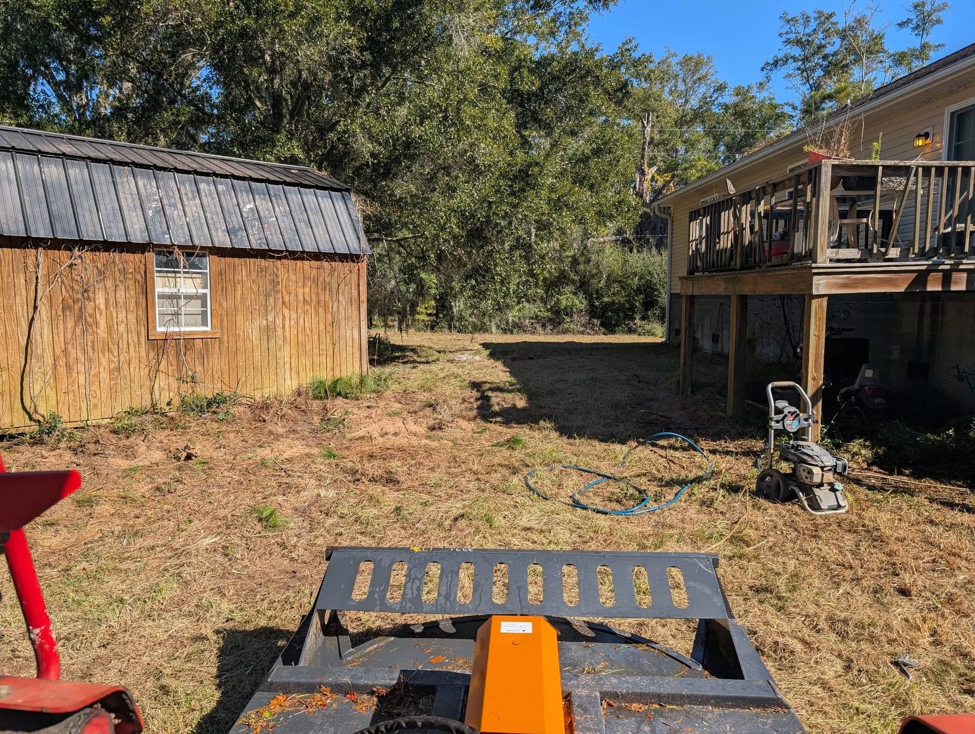 Backyard with a wooden shed, raised deck, and leaf blower in the foreground on dry ground.