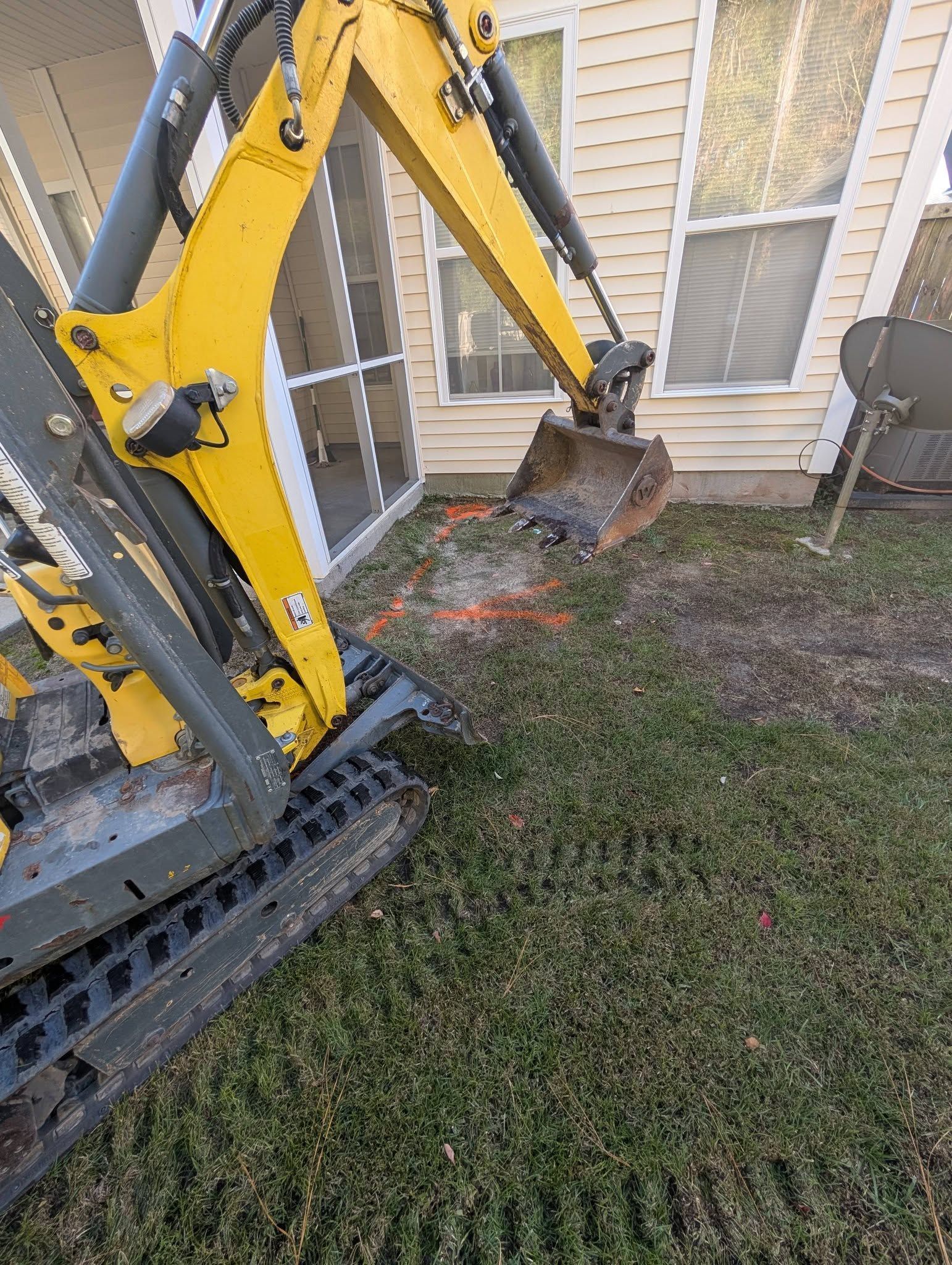 Yellow excavator bucket digging grass beside a house wall and window.