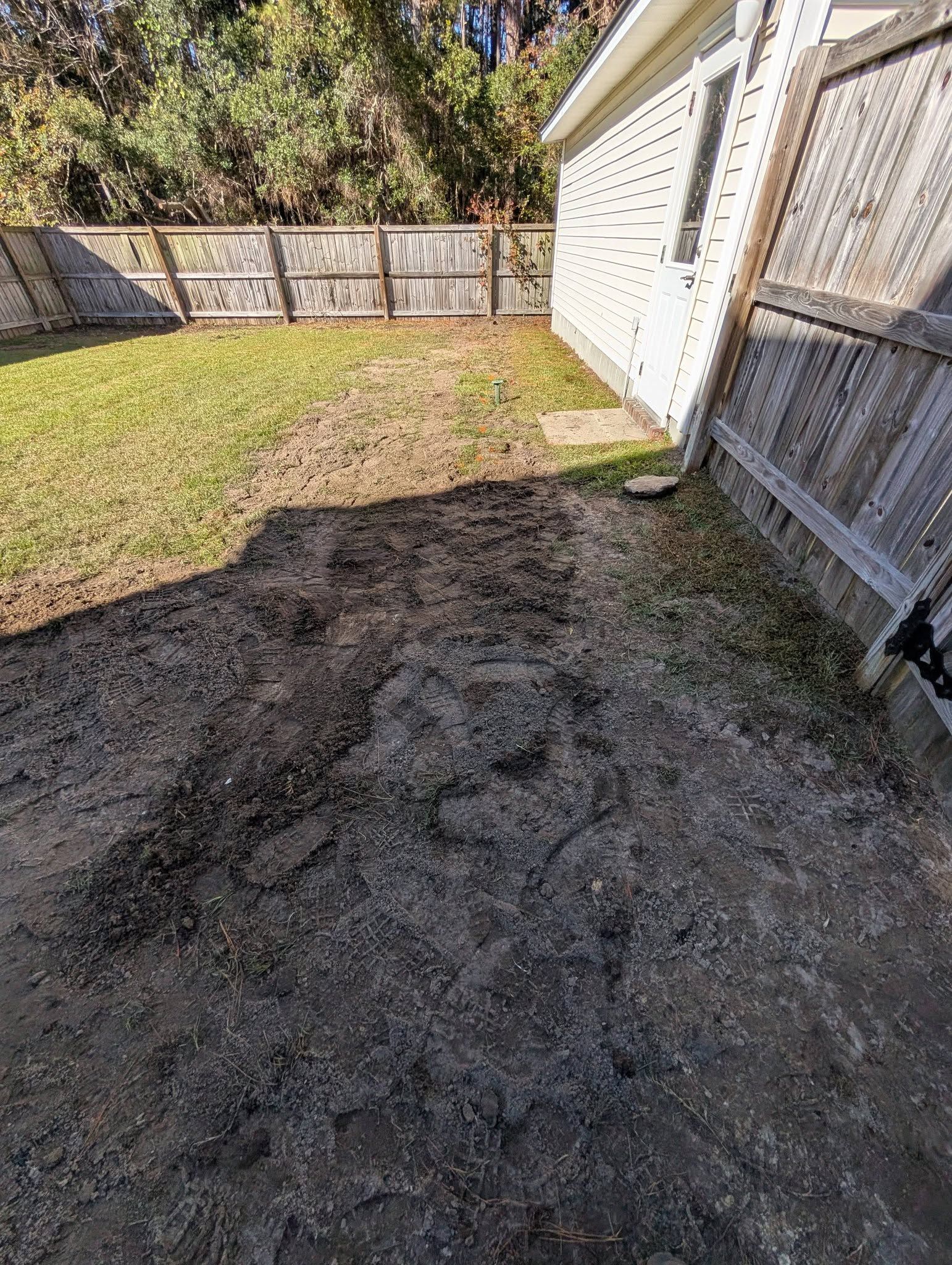 Backyard with a bare dirt patch beside a house and fenced lawn in bright sunlight