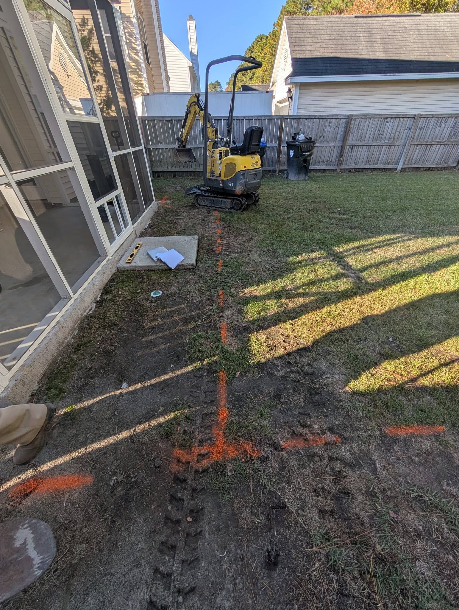 Backyard lawn with marked trench line, excavator, and house wall beside a screened porch.