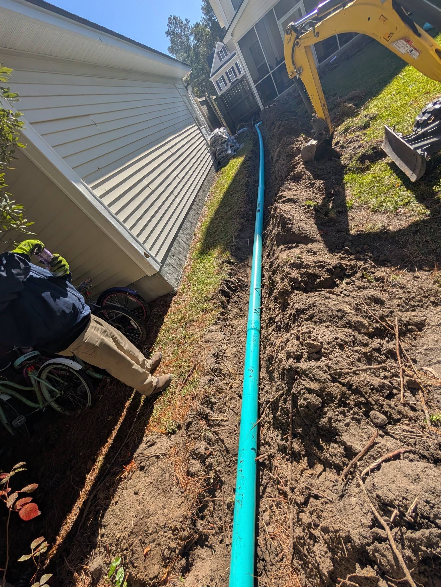 Backyard trench beside a house with a blue pipe and yellow excavator on a sunny day