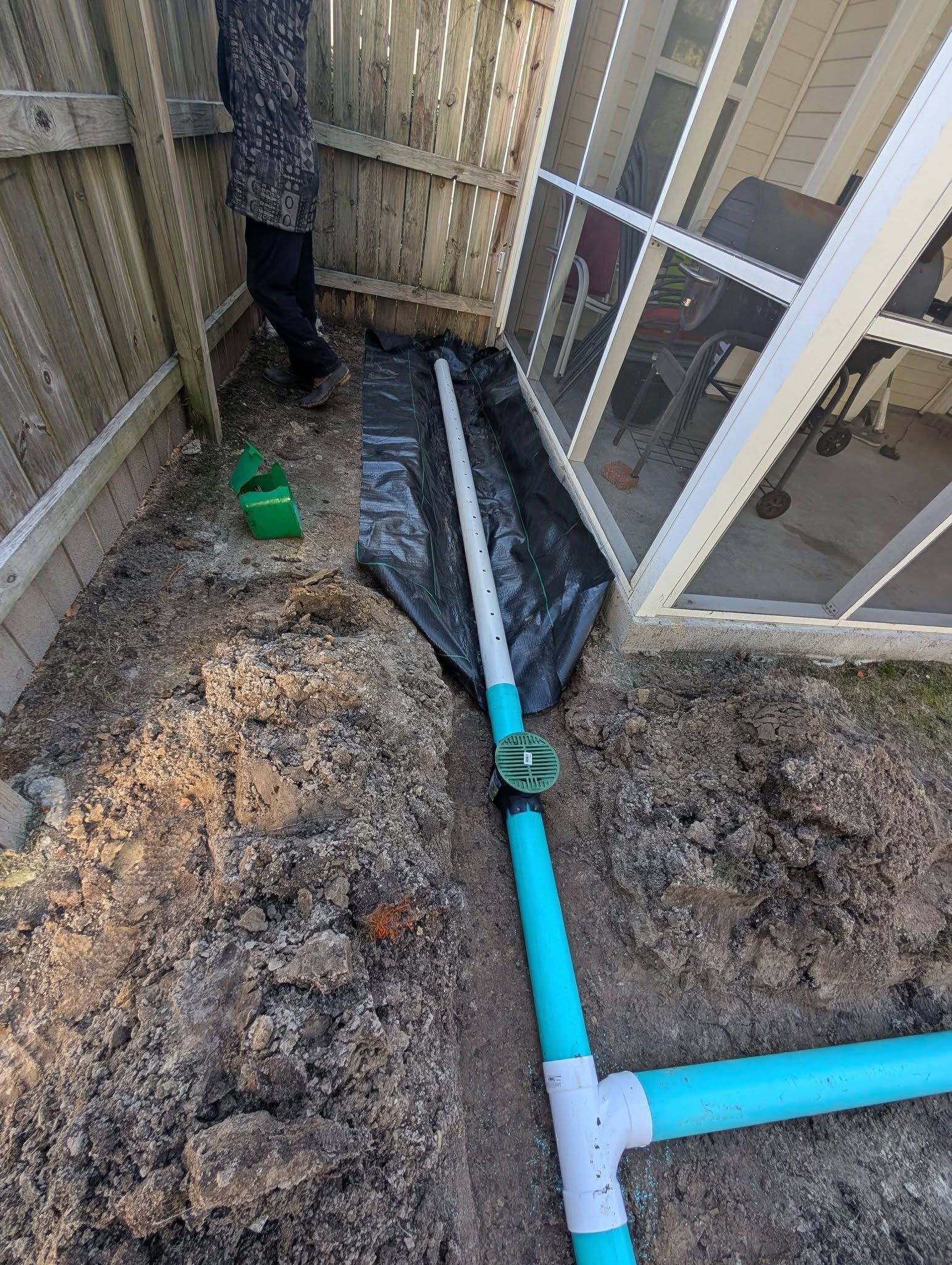 Outdoor trench with blue PVC pipes and white fittings beside a fenced structure.
